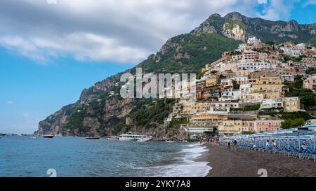 I visitatori passeggiano lungo la spiaggia di ciottoli di Positano, circondata da colorate case sulla scogliera sotto un cielo azzurro. Foto Stock