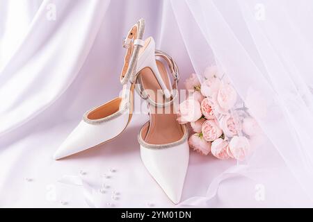 Elegante composizione per matrimoni con eleganti scarpe bianche, bouquet di peonie rosa su fondo in raso con velo bianco. natura morta. sposa mattutina Foto Stock