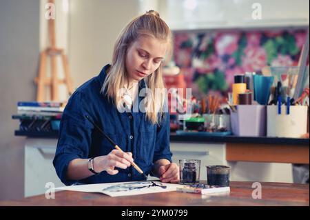 Giovane artista che crea un dipinto ad acquerello in uno studio luminoso e colorato pieno di materiali d'arte Foto Stock