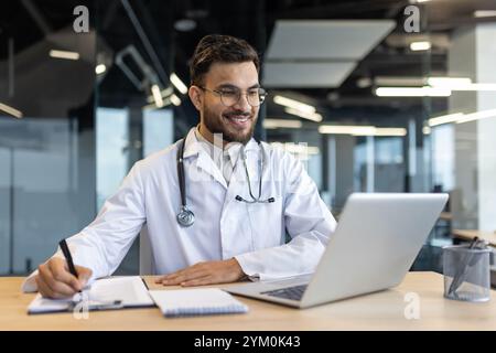 Un medico che indossa uno stetoscopio si occupa di consulenze online sul laptop. Sorridere, prendere appunti, multitasking in un ambiente d'ufficio moderno. Esperienza professionale ed efficiente di telemedicina. Foto Stock