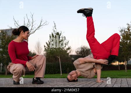 Un uomo con pantaloni rossi in posa breakdance in un parco, mentre una donna lo guarda. La scena è informale e rilassata, con l'uomo e la donna che sorridono e si divertono Foto Stock