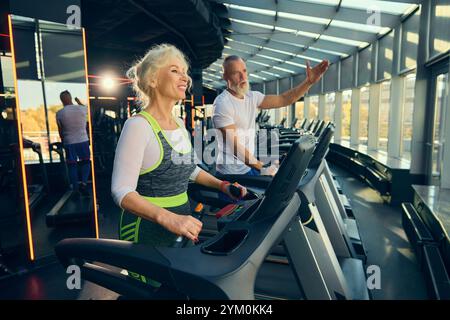 Uomo e donna anziani, coppia impegnata in un allenamento con tapis roulant in una vivace palestra. Motivazione a rimanere attivi e sani. Foto Stock