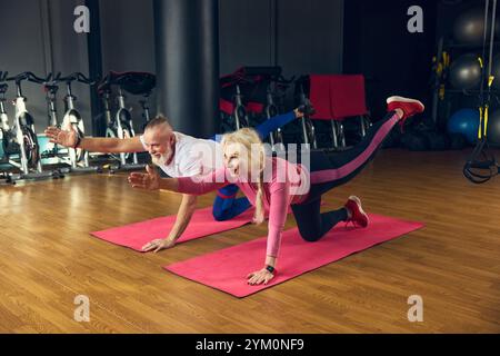 Uomo e donna anziani in equilibrio su un ginocchio con un braccio allungato sui tappetini da yoga in palestra, in allenamento, promuovendo coordinazione e forza. Foto Stock