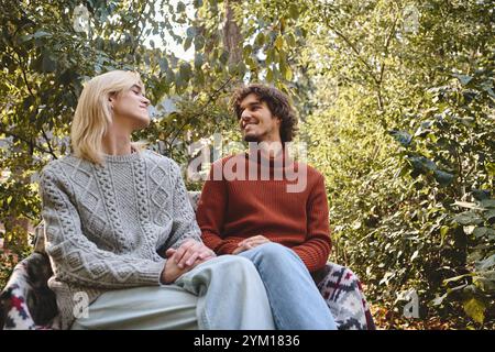 Due giovani e belle persone condividono una connessione gioiosa in un ambiente all'aperto vibrante. Vestiti in modo caldo, sono comodamente seduti in una lussureggiante vegetazione, radiosa Foto Stock