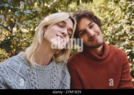 Giovani coppie gay amanti si godono compagnia a vicenda in un lussureggiante giardino pieno di verde. Indossano maglioni confortevoli e condividono sorrisi caldi, catturando un mese Foto Stock