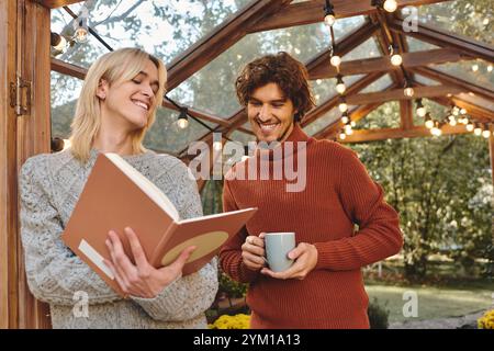 Giovani coppie gay che amano sorridono mentre si impegnano con un libro in un affascinante spazio all'aperto adornato di luci. L'atmosfera irradia calore e connessione Foto Stock