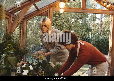 Giovani coppie gay amanti si impegnano nel giardinaggio all'interno di un'incantevole serra, circondata da una vegetazione vivace. Si concentrano sulla nutrizione di piante delicate Foto Stock