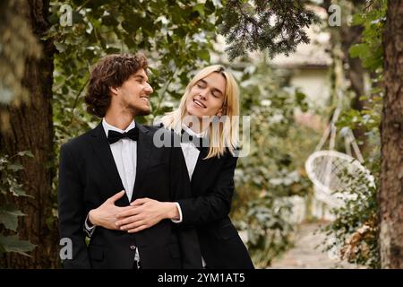 Due bellissimi giovani uomini si abbracciano giocosamente, vestiti con eleganti abiti neri con lacci, circondati da una vegetazione lussureggiante. Foto Stock