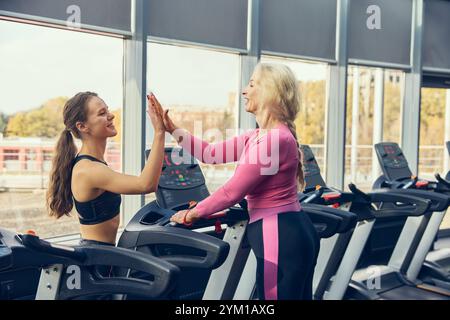 Una donna anziana e il suo fitness trainer condividono cinque punti sul tapis roulant, celebrando il progresso e la motivazione del fitness. Foto Stock