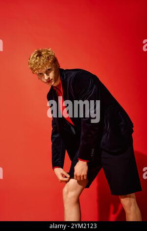 Un uomo elegante sfoggia uno stile unico per le vacanze in un vivace ambiente rosso. Combina un blazer scuro con pantaloncini corti, che irradiano fiducia e creatività Foto Stock