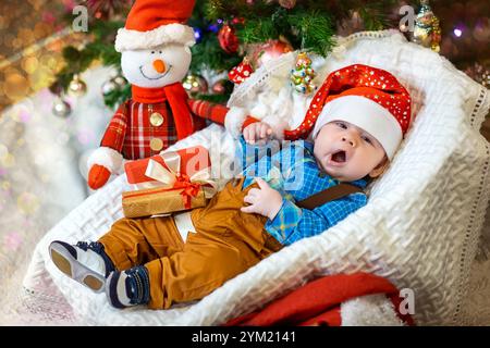 Un bambino sonnolento indossa un cappello di Babbo Natale e un abbigliamento accogliente, annidato da decorazioni natalizie e da un allegro pupazzo di neve. Foto Stock