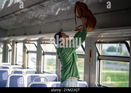 Man Tourist solleva lo zaino sul portabagagli sospeso in treno. Comodità di trasporto e trasporto in solitaria Foto Stock