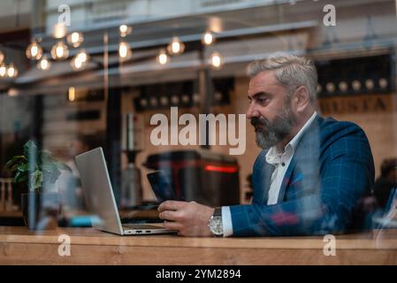Un uomo d'affari maturo che usa il telefono mentre lavora su un portatile in ufficio vicino alla finestra. Foto Stock