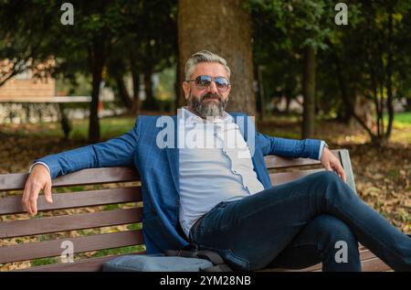 Un uomo d'affari maturo e felice si rilassa sedendosi sulla panchina del parco. Foto Stock