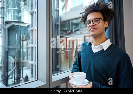 17 novembre 2024: Un giovane con i bicchieri e un maglione riflette tranquillamente con una tazza di caffè, possibilmente su un progetto di apprendimento o un'idea. Immagine simbolica *** Ein junger Mann mit Brille und Pullover reflektiert in Ruhe mit einer Tasse Kaffee, möglicherweise über ein Lernprojekt oder eine idee. Symbolbild Foto Stock