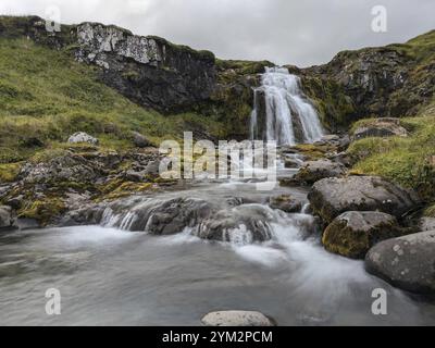 Cascata che si estende su rocce muschiate con acqua corrente e vegetazione sotto un cielo nuvoloso. Islanda Foto Stock
