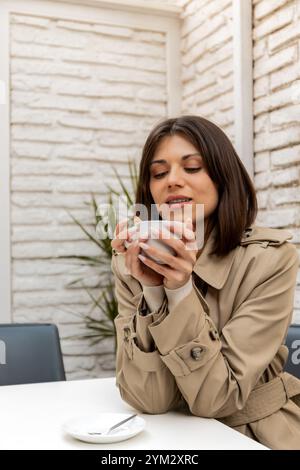 Una giovane donna con un trench beige regala una tazza calda, assaporando un momento di pace in un caffè all'aperto. Lo sfondo neutro e l'illuminazione soffusa creano un effetto Foto Stock