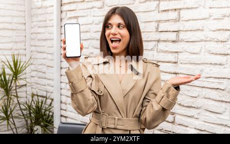Una giovane donna gioiosa in un trench beige regge uno smartphone con uno schermo vuoto, esprimendo emozioni. Perfetto per promozioni su app o prodotti digitali Foto Stock
