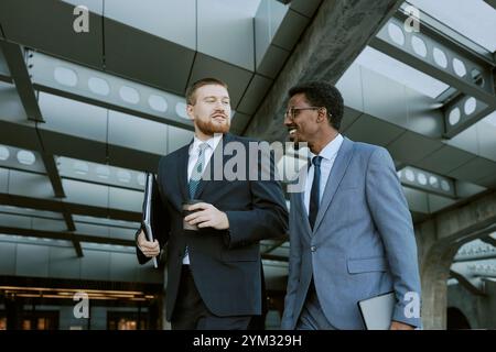 Due uomini d'affari, uno caucasico e uno afro-americano, si impegnano in conversazione mentre si trovano all'aperto in un edificio di uffici, entrambi in possesso di documenti e vestiti con abiti Foto Stock