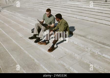 Due uomini seduti su gradini di pietra all'aperto, che collaborano e discutono di qualcosa sullo schermo di un notebook. Colleghi multirazziali che lavorano e condividono idee in ambiente urbano Foto Stock