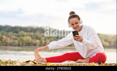 Donna che indossa le cuffie wireless, sorride e utilizza il suo smartphone mentre è seduta su un tappetino da yoga e fa esercizi di stretching all'aperto vicino ai riversi Foto Stock