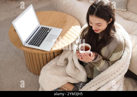 Giovane donna con tazza di tè e laptop a casa il giorno d'inverno Foto Stock