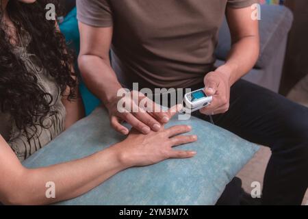 Primo piano di un uomo misura il polso di una donna e la saturazione di ossigeno con un pulsossimetro seduto sul divano a casa, concetto sanitario, pulsossimete Foto Stock