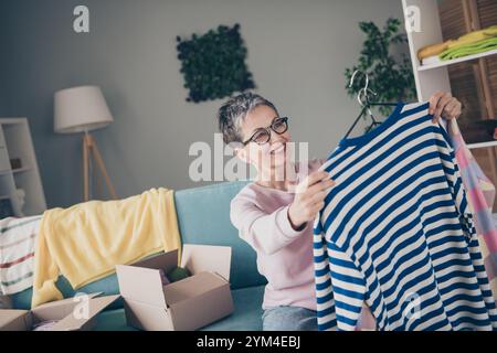 Foto di adorabile dolce signora indossa una felpa rosa scegliendo abiti da donazione all'interno della casa della stanza Foto Stock