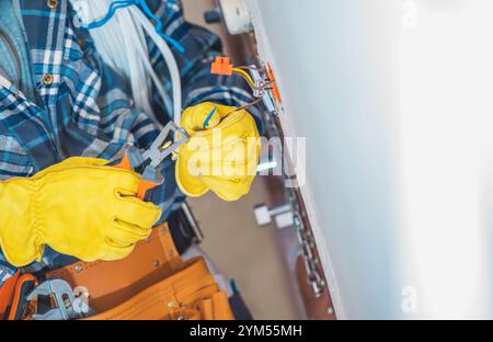 Un elettricista con guanti gialli utilizza attrezzi per lavorare sul cablaggio elettrico in un'area residenziale. L'attività si svolge in un ambiente interno luminoso. Foto Stock