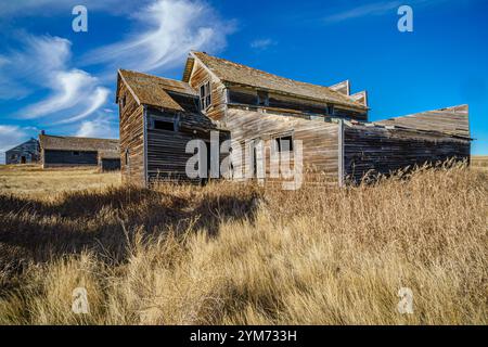 Una grande e vecchia casa si trova in un campo di erba alta. La casa è circondata da altri vecchi edifici, e il cielo è limpido e blu. La scena è tranquilla e.. Foto Stock