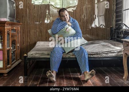 Un uomo solitario in pigiama siede su un materasso e abbraccia un cuscino Foto Stock