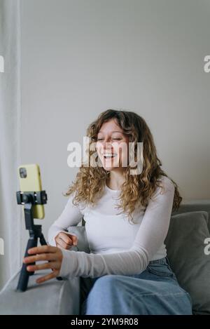 Giovane donna con i capelli ricci che ride mentre registra un video blog utilizzando uno smartphone su un cavalletto, creando contenuti coinvolgenti per il suo pubblico online Foto Stock
