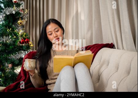 Una rilassata donna asiatica in abiti caldi e accoglienti legge un libro e si gode una bevanda calda sul divano nel soggiorno, immergendosi nell'atmosfera tranquilla di Foto Stock