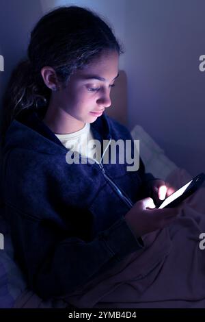 Teenager triste e stanca che usa uno smartphone sdraiato a letto a tarda notte, gioca, guarda video e digita. Dipendenza da schermo per bambini. SC Foto Stock