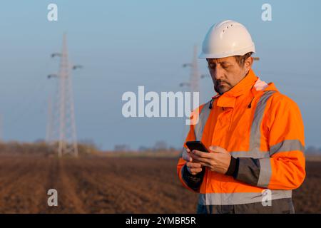 Ingegnere elettrico che utilizza lo smartphone sul campo vicino a piloni elettrici, messa a fuoco selettiva Foto Stock