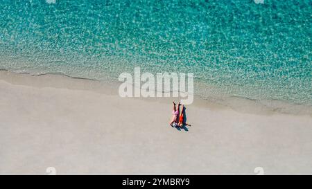 La vista aerea di una giovane coppia sposata in costume da bagno si trova sulla riva dell'oceano. Un uomo e una donna con i cappelli estivi si rilassano sulla spiaggia. Marito An Foto Stock