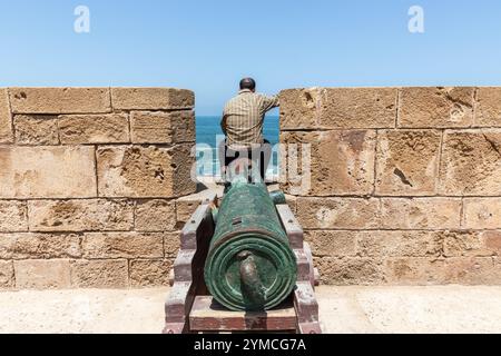 Arroccato su un vecchio cannone nella fortezza di Essaouira, un uomo contempla il mare Foto Stock