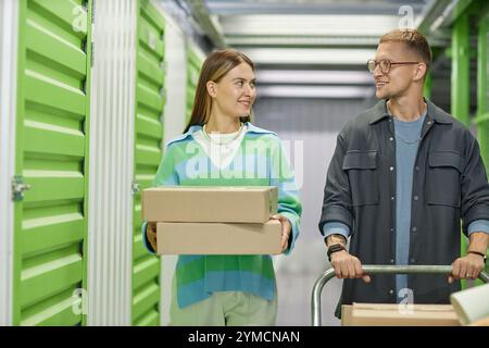 Foto media di donna sorridente che trasporta scatole di cartone che cammina lungo il corridoio del magazzino con l'aiuto del fidanzato che spinge il carrello Dolly caricato Foto Stock