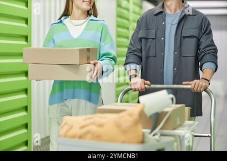 Colpo ritagliato di donna sorridente che porta una pila di scatole di cartone che cammina accanto al fidanzato aiutando a spostarsi, mentre spinge il carrello Dolly caricato in h Foto Stock