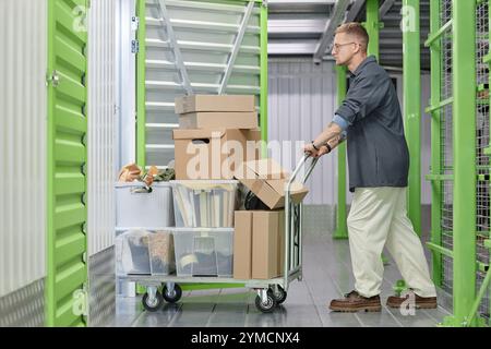 Foto a tutta lunghezza di un bell'uomo che carica il carrello con scatole di cartone in un'unità di immagazzinamento del magazzino mentre si sposta o si rifiuta l'appartamento Foto Stock