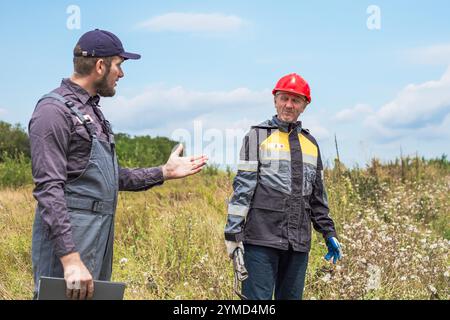 Un appaltatore e un lavoratore in tuta con palanchino e chiave regolabile in un'area rurale discutono del lavoro. Lavoratori municipali, meccanici e idraulici. Foto Stock