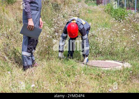 Un appaltatore e un lavoratore in tute con palanchino e chiave regolabile in un'area rurale aprono un pozzo per riparare una rete idrica. Lavoratori municipali. Foto Stock