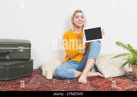 Donna caucasica con capelli biondi in un maglione giallo e jeans sorride tenendo in mano una tavoletta vuota. Seduti su un tappeto decorativo con piante e valica Foto Stock