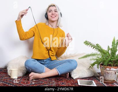 Donna caucasica con capelli biondi, indossa un maglione giallo e cuffie, seduta a gambe incrociate su un tappeto, sorridente, con una foglia di felce all'interno. Foto Stock
