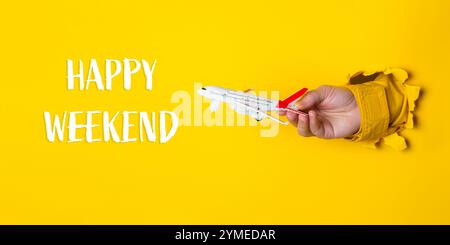 Mano che regge un aereo giocattolo davanti a uno sfondo giallo con le parole Happy Weekend scritte di seguito Foto Stock