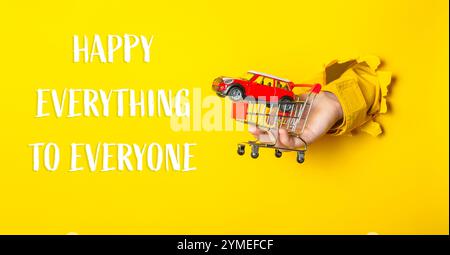 Mano che tiene un'auto giocattolo su un carrello della spesa con le parole "Happy Everything" (felice tutto a tutti) scritte qui sotto Foto Stock