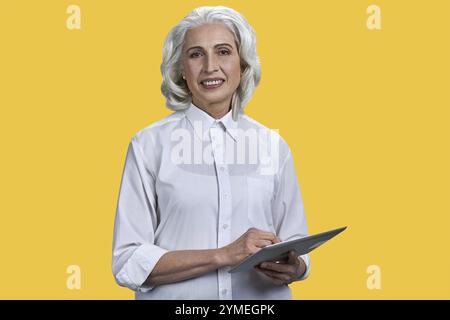 Donna d'affari matura sorridente che tiene in mano un pc un tablet e guarda la fotocamera. Bella donna anziana con tablet pc digitale su sfondo giallo Foto Stock