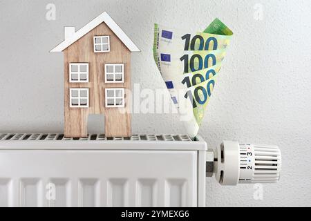 Casa modello in legno accanto a centinaia di euro di bollette su radiatore con termostato, radiatore divora euro di denaro, simbolo dell'aumento dei costi di riscaldamento Foto Stock