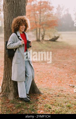 Una giovane donna con i capelli ricci si appoggia contro un albero robusto, tenendo una bevanda calda. Vestita con un comodo cappotto grigio, gode dell'atmosfera tranquilla di un Foto Stock