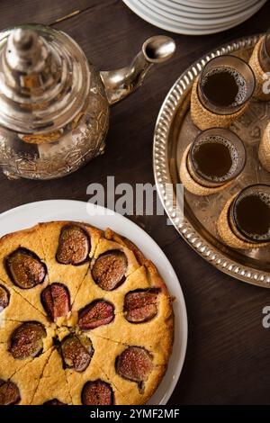 Tradizionale tè marocchino con torta di fichi servito su un rustico tavolo di legno, che mette in mostra dettagli decorati, autenticità culturale e dolce indulgenza. Foto Stock
